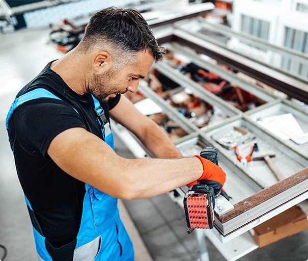 Handyman sanding a wooden window frame using a power sander in a workshop.